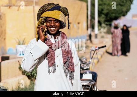 Entzückte ägyptischen Mann in Turban in der Straße an sonnigen Tag stehen und mit einem Anruf, während Blick auf die Kamera Stockfoto