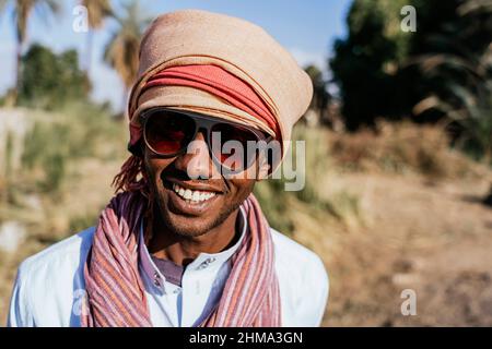 Fröhlicher arabischer Mann in Sonnenbrille mit traditionellem Turban, der die Kamera anschaut und lächelt, während er auf dem verschwommenen Hintergrund einer exotischen Stadt in Summe steht Stockfoto