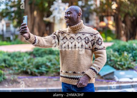 Fröhlicher afroamerikanischer Mann in warmem Strickpullover und Jeans, der Selfie-Fotos auf dem Handy fotografiert, während er an sonnigen Tagen im Park steht Stockfoto