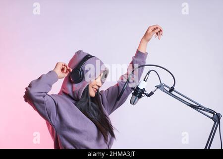 Junge talentierte Frau mit Hoodie und Kopfhörern tanzt und singt ins Mikrofon mit Pop-Filter, während sie im Modus Lied auf purpurem Hintergrund aufzeichnet Stockfoto