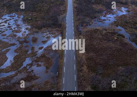 Drone Blick auf lange schmale Asphaltstraße mit Herstellung Linien zwischen Feuchtgebiet mit verdorrtem Gras in der Natur von Norwegen laufen Stockfoto