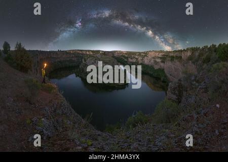 Seitenansicht eines nicht erkennbaren männlichen Touristen mit Fackel, die Berge bewundern, die sich in der Nacht unter dem Sternenhimmel im Wasser spiegeln, felsige Küste mit Bäumen gegen Dunkelheit Stockfoto