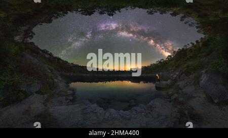 Seitenansicht eines nicht erkennbaren männlichen Touristen mit Fackel, die Berge bewundern, die sich in der Nacht unter dem Sternenhimmel im Wasser spiegeln, felsige Küste mit Bäumen gegen Dunkelheit Stockfoto