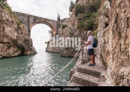 Seitenansicht eines reifen Mannes mit grauem Haar, der am Ufer in der Nähe des plätschernden Meeres gegen eine alte Brücke in bergigem Gelände steht Stockfoto