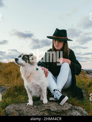 Ganzer Körper von stilvollen Hündin in schwarzem Hut sitzt auf grasbewachsenen Boden mit niedlichen weißen australischen Schäferhund in der Natur Stockfoto