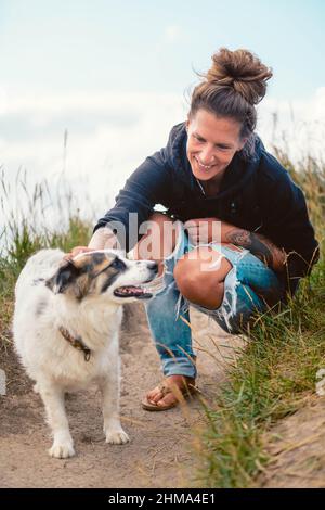 Positive weibliche Besitzerin sitzt auf Hacken und streicheln gehorsame Border Collie auf dem Weg während des Spaziergangs auf einer Wiese in Kantabrien Stockfoto