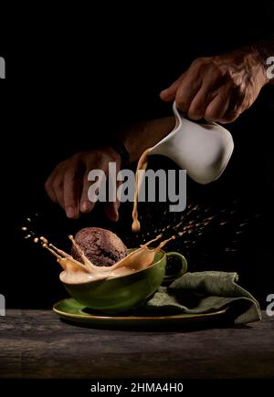 Crop anonyme Person wirft leckere süße Kekse in grüne Keramik-Tasse mit Spritzkaffee auf dem Tisch in der Küche auf schwarzem Hintergrund Stockfoto