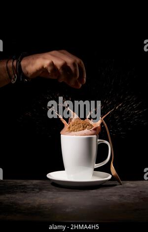 Crop anonyme Person wirft leckere süße Kekse in weiße Keramik-Tasse mit Spritzkaffee auf dem Tisch in der Küche auf schwarzem Hintergrund Stockfoto