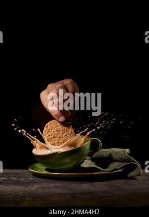 Crop anonyme Person wirft leckere süße Kekse in grüne Keramik-Tasse mit Spritzkaffee auf dem Tisch in der Küche auf schwarzem Hintergrund Stockfoto