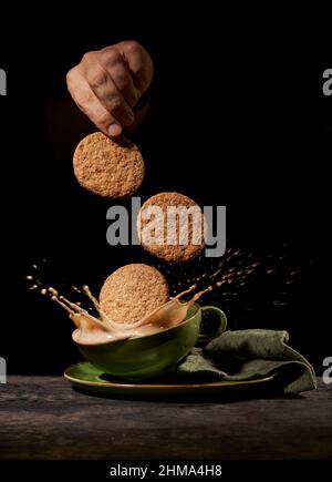 Crop anonyme Person wirft leckere süße Kekse in grüne Keramik-Tasse mit Spritzkaffee auf dem Tisch in der Küche auf schwarzem Hintergrund Stockfoto