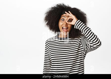 Aufgeregt junge afrikanische Frau zeigt okay, Null Geste, ok Zeichen über Auge und lächelnd erstaunt, Auschecken smth genial, sieht beeindruckt, weiß Stockfoto