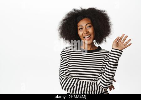 Freundliche afroamerikanische Frau, die hallo sagt, die Hand zur Kamera schwenkt, Sie begrüßt, Sie begrüßt, Sie begrüßt, Sie lächelt sorglos vor der Kamera, steht auf weißem Hintergrund Stockfoto