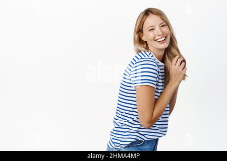 Schönheit. Attraktive blonde natürliche Frau lachend, berührt ihre Haare und lächelt kokett an der Kamera, stehend in T-Shirt und Jeans über weiß Stockfoto