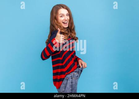 Seitenansicht einer extrem glücklichen Frau, die einen gestreiften Pullover im lässigen Stil trägt, den Daumen nach oben zeigt und sich mit einer erfolgreichen Gewichtsabnahme und einer Diät rühmt. Innenaufnahme des Studios isoliert auf blauem Hintergrund. Stockfoto