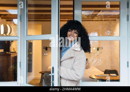 Fröhliche junge afroamerikanische Frau mit lockigen dunklen Haaren in warmem Mantel, die durch eine Glastür ins Haus kommt und wegschaut Stockfoto
