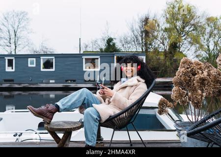 Entzückte junge afroamerikanische Hündin in warmem Mantel mit Smartphone und Musik in Kopfhörern, während sie am See chillte Stockfoto