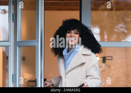 Fröhliche junge afroamerikanische Frau mit lockigen dunklen Haaren in einem warmen Mantel, die durch die Glastür aus dem Haus geht und die Kamera anschaut Stockfoto