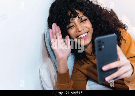 Entzückte junge afroamerikanische Frau mit lockigen dunklen Haaren, die auf dem Smartphone per Videoanruf sprach, während sie sich zu Hause auf dem Bett ausruhte Stockfoto