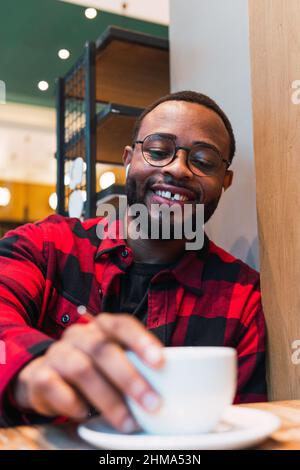 Von unten ein glücklicher bärtiger afroamerikanischer Mann im karierten Hemd mit Ohrhörern, der heißes Getränk aus der Tasse trank, während er sich im Café ausruhte und nach d schaute Stockfoto