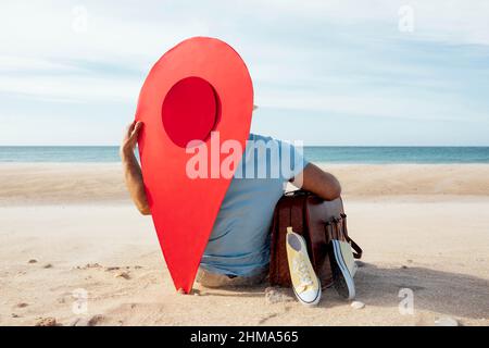 Rückansicht eines nicht erkennbaren männlichen Touristen, der im Sommer mit Gepäck und rotem Kartenzeiger an der sandigen Küste sitzt Stockfoto