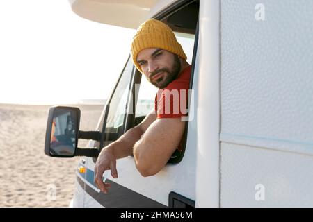 Männlicher Reisender, der in einem Reisewagen am Meer geparkt ist und die Kamera anschaut Stockfoto