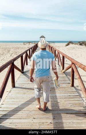 Blick auf den Rücken eines Mannes, der an sonnigen Sommertagen barfuß auf einer hölzernen Promenade am sandigen Ufer zum Meer läuft Stockfoto