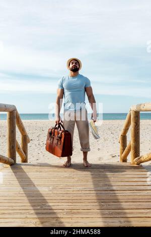Ganzer Körper von männlichen Touristen stehen mit Gepäck und Gummischuhen auf Holzboardwalk an der Küste während der Sommerferien und Blick auf die Kamera Stockfoto