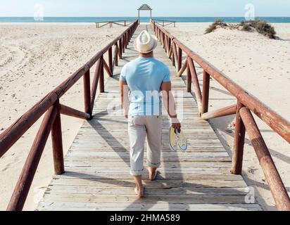 Blick auf den Rücken eines Mannes, der an sonnigen Sommertagen barfuß auf einer hölzernen Promenade am sandigen Ufer zum Meer läuft Stockfoto