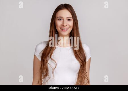 Porträt einer schönen Frau mit gutherzigen Blick und charmantem Lächeln, stehend und auf die Kamera schauend, positive Emotionen ausdrückend, trägt weißes T-Shirt. Innenaufnahme des Studios isoliert auf grauem Hintergrund. Stockfoto