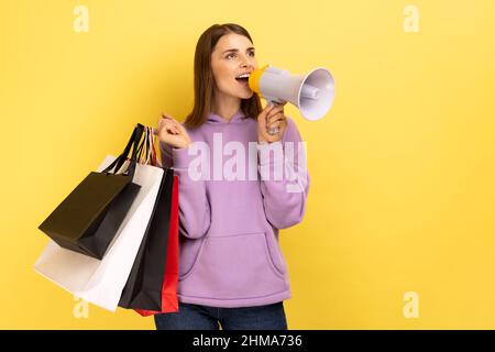 Junge schöne Frau mit braunen welligen Haaren hält Einkaufstaschen und Megaphon, kündigt Verkauf und Rabatt, trägt lila Hoodie. Innenaufnahme des Studios isoliert auf gelbem Hintergrund. Stockfoto