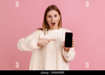 Porträt einer staunenden Frau, die das Mobiltelefon in den Händen hält, auf das leere Display des Smartphones mit Platz für die Werbung zeigt und einen weißen Pullover trägt. Innenaufnahme des Studios isoliert auf rosa Hintergrund. Stockfoto