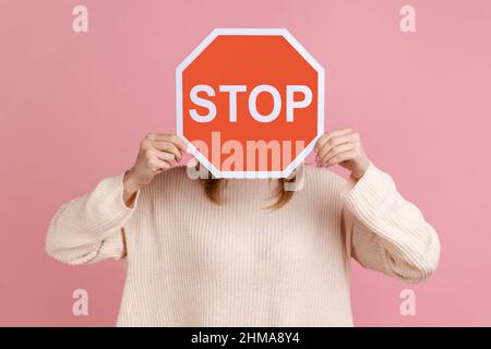 Porträt einer unkenntlichen Frau, die ihr Gesicht mit rotem Schild versteckt, Kampagne gegen häusliche Gewalt, Schutz der Frauenrechte, in weißem Pullover. Innenaufnahme des Studios isoliert auf rosa Hintergrund. Stockfoto