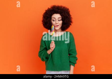 Hungrige lustige Frau mit Afro-Frisur trägt grünen lässigen Stil Pullover mit bunten Süßigkeiten, lecken, Blick auf süß mit kindischem Ausdruck. Innenaufnahme des Studios isoliert auf orangefarbenem Hintergrund. Stockfoto