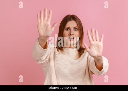 Porträt einer verängstigten jungen erwachsenen blonden Frau, die mit Handflächen zum Stoping aufsteht und mit verängstigten Augen überrascht aussieht und einen weißen Pullover trägt. Innenaufnahme des Studios isoliert auf rosa Hintergrund. Stockfoto