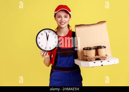 Porträt einer lächelnden Kurierin, die Wanduhr und Kaffee mit Pizzakarton hält, Express-Lieferung rechtzeitig, Overalls und rote Mütze trägt. Innenaufnahme des Studios isoliert auf gelbem Hintergrund. Stockfoto