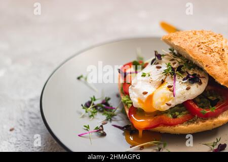 Pochiertes Ei auf einem Brötchen mit Avocado, Paprika und Mikrogrün auf einem grauen Teller. Gesundes Frühstück. Brunch-Idee. Sauberes Essen. Vegetarisch. Eiweißsauer Stockfoto