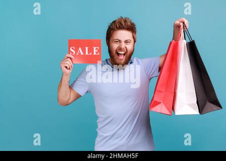 Porträt eines aufgeregten bärtigen Mannes, der Papiertüten und Verkaufsschild hält, Einkaufen und Rabatte, die Kamera anschaut und fröhlich schreit. Innenaufnahme des Studios isoliert auf blauem Hintergrund. Stockfoto