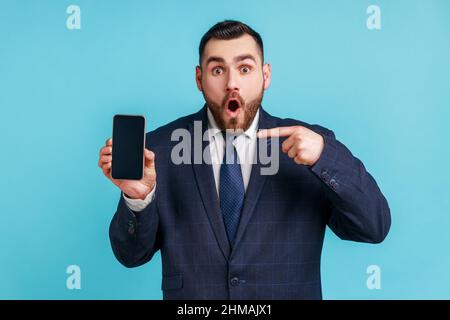 Überraschender bärtiger Mann mit offiziellem Anzug, der mit dem Finger auf das Smartphone mit weißem, leeren Display zeigt, schockiert über neue Telefonfunktionen. Innenaufnahme des Studios isoliert auf blauem Hintergrund. Stockfoto