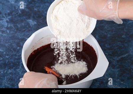 Mehl wird der Schokoladenmasse hinzugefügt. Der Prozess der Herstellung eines Käse-Brownie. Blauer Marmorhintergrund. Nahaufnahme. Stockfoto