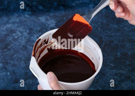 Heiße Schokolade mit einem Silikonspachtel umrühren. Dieser Prozess findet in einer weißen Schale statt. Blauer Marmorhintergrund. Stockfoto