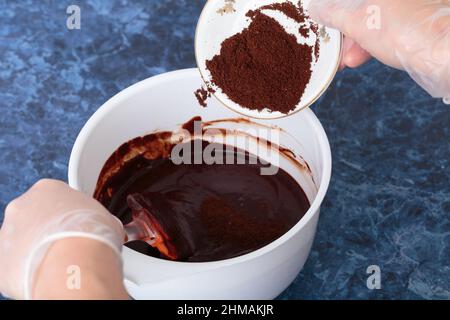 Hinzufügen von grobem Kaffee zu heißer Schokolade. Dieser Prozess findet in einer weißen Schale statt. Blauer Marmorhintergrund. Stockfoto