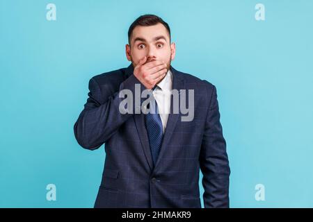 Ich werde es nicht sagen. Porträt eines staunenden Geschäftsmannes, der einen offiziellen Anzug trägt, der den Mund mit der Hand bedeckt, geheim hält, erschrocken vor schockierenden Nachrichten. Innenaufnahme des Studios isoliert auf blauem Hintergrund. Stockfoto