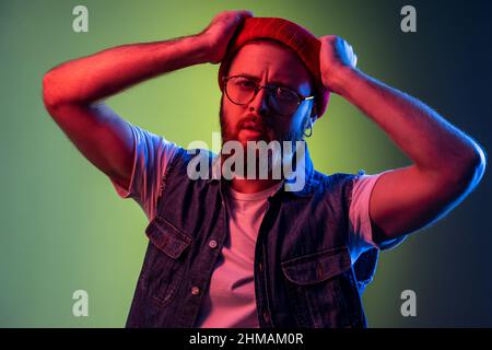 Junger erwachsener bärtiger Hipster mit Brille, Blick auf die Kamera, erhobene Arme, Ausdruck nachdenklicher verträumter Emotionen, trägt roten Beanie-Hut. Innenaufnahmen im Studio, isoliert auf farbigem Neonlicht-Hintergrund. Stockfoto