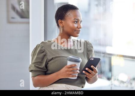 Das Internet ist mein Bindeglied zur Welt da draußen. Aufnahme einer Geschäftsfrau, die Kaffee trinkt und ihr Handy benutzt, während sie aus ihrem Bürofenster schaut. Stockfoto