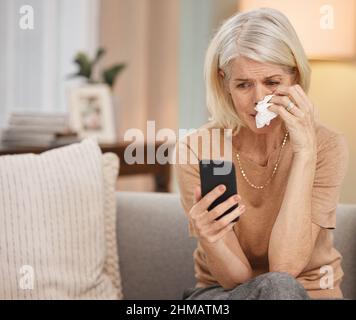 2020, das Jahr des Traumas. Aufnahme einer reifen Frau, die ein Smartphone benutzt und zu Hause auf dem Sofa weint. Stockfoto