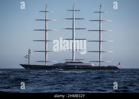 An sonnigen Tagen steht in der Bucht bei Monaco eine riesige Segelyacht in konzeptuellem Design mit drei Masten, die Segel werden abgesenkt, die Yacht dreht sich um sie Stockfoto