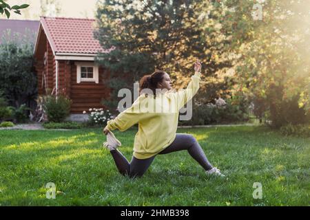Kaukasische übergewichtige Frau in Sportkleidung, die auf grünem Gras mit Holzhaus und hohen Bäumen im Hinterhof körperliche Übungen macht Stockfoto