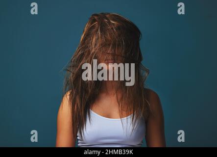 Es ist alles chaotisch. Die Haare, das Bett, die Worte, sogar das Herz. Studioaufnahme einer jungen Frau, die ihr Gesicht mit ihren Haaren vor blauem Hintergrund bedeckt. Stockfoto