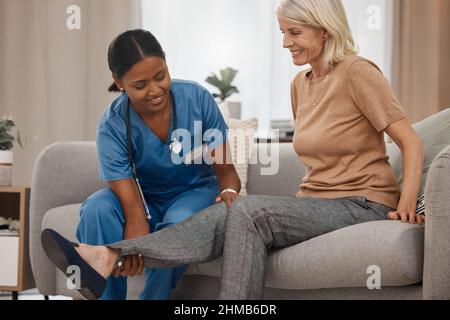 Eine helfende Hand kann viel heilen. Aufnahme eines Arztes, der zu Hause eine ältere Frau am Knöchel auf dem Sofa untersucht. Stockfoto