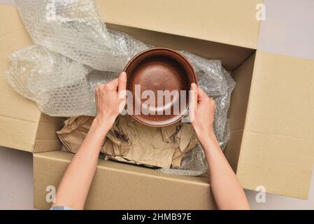 Frau packt Geschirr in eine Schachtel mit Geschenkpapier und Filmansicht von oben. Umzug in ein neues Haus. Auspacken von Waren. Stockfoto